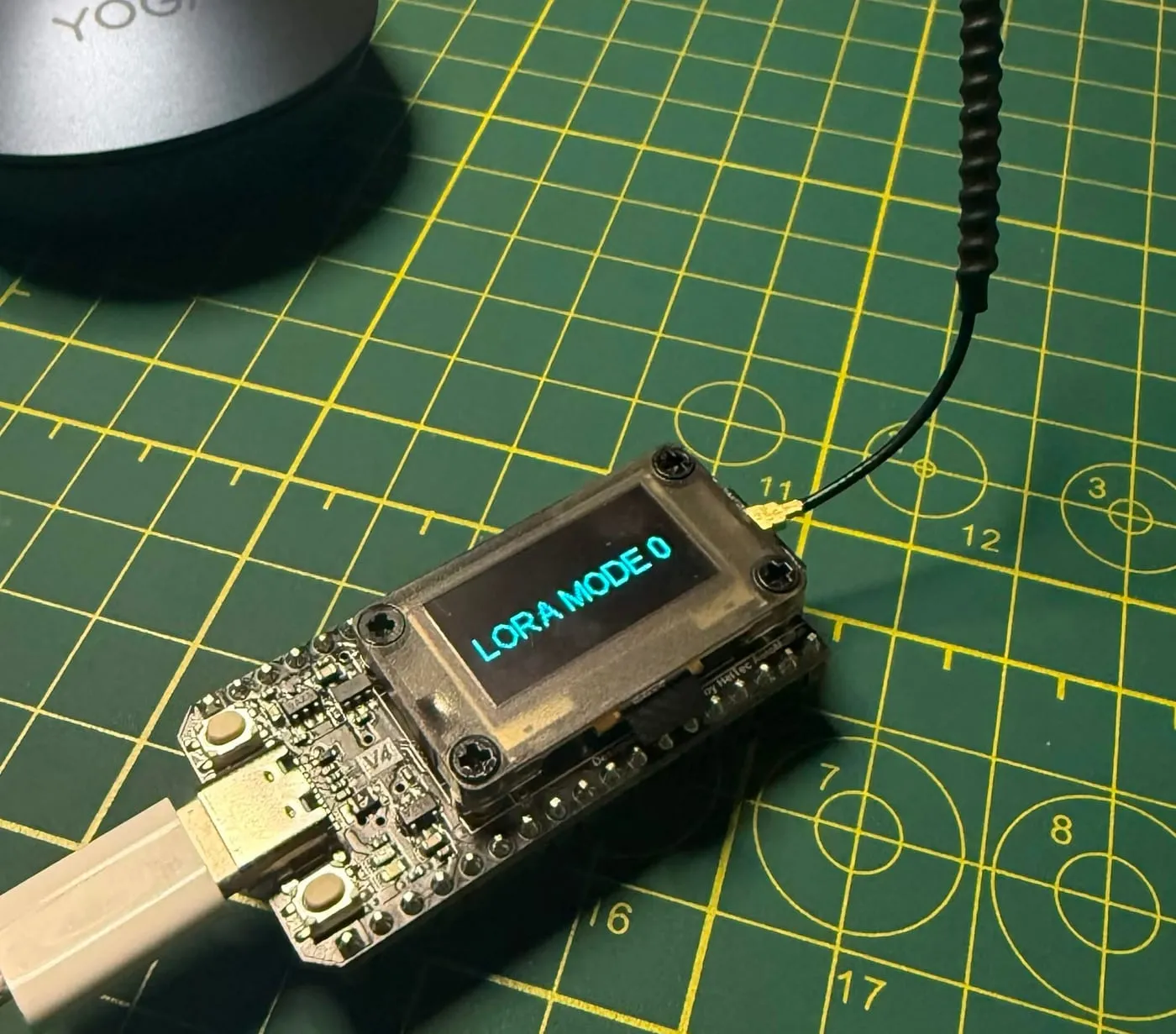 a heltec wifi lora 32 v4 board on a green cutting mat with its spring antenna attached and its oled reading lora mode 0, plugged in over usb-c